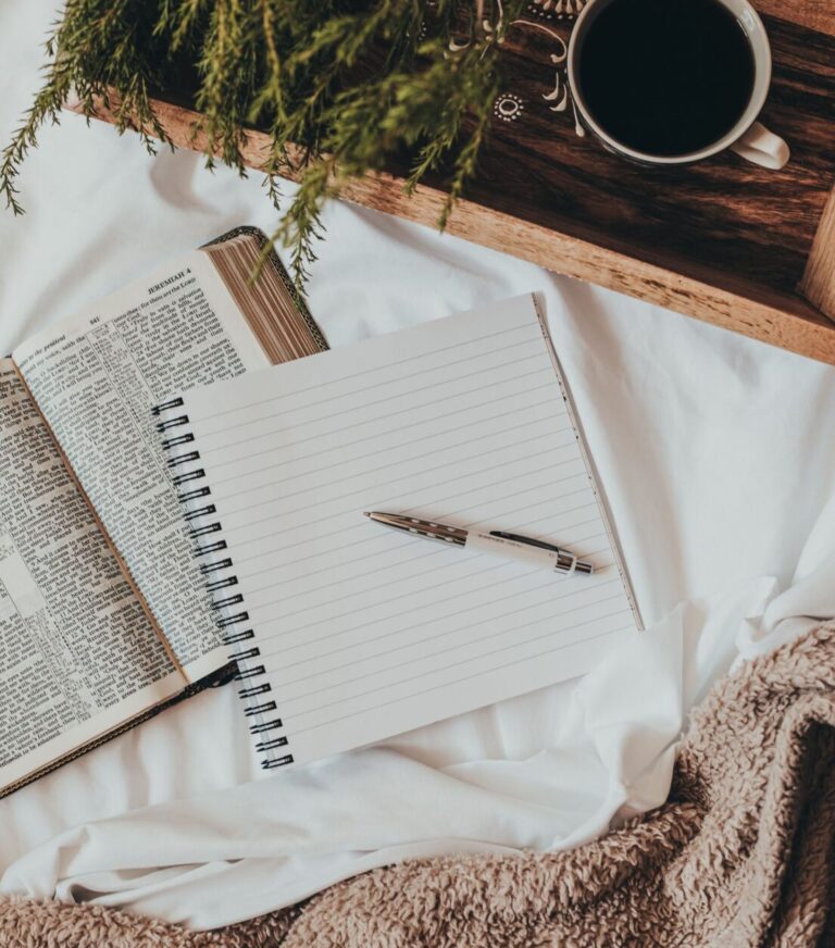 Bible, pen, notebook, and tray with coffee sitting atop a white sheet with brown fuzzy blanket to the side