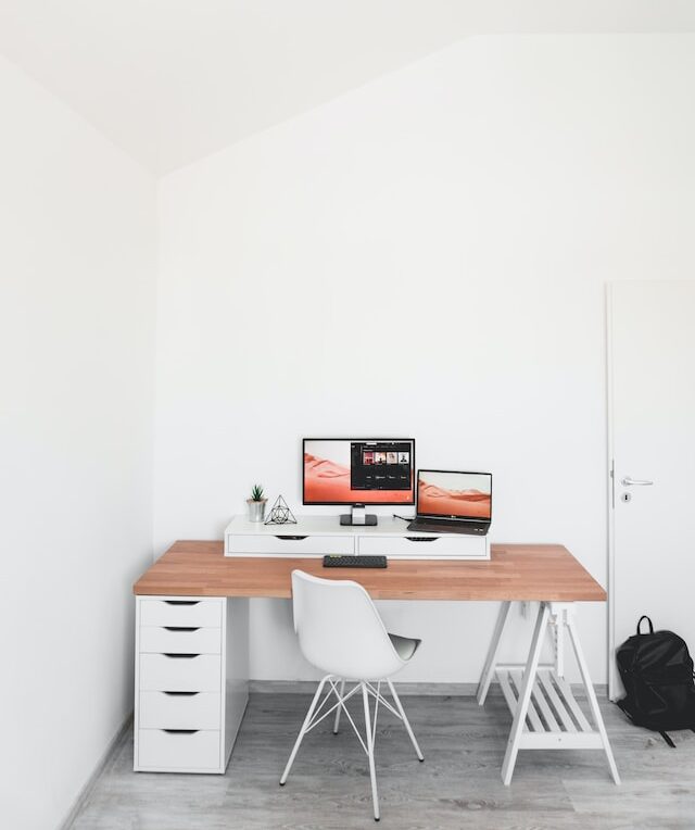 A white room with a singular workspace. A white desk with a natural wood desktop and white share. Two computer monitors display a well-maintained website.